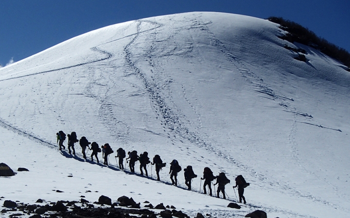 A group of people wearing backpacks hike in a line up a snowy incline.